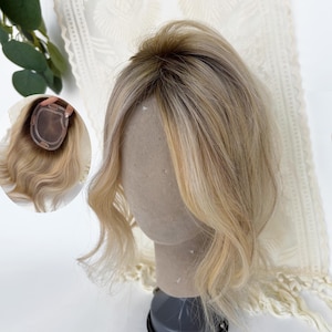 May include: A hair topper featuring wavy, shoulder-length blonde hair with darker roots. The hair is styled with a centre part and soft waves. An inset circular image shows the underside of the hair topper, revealing a sheer base with clips.