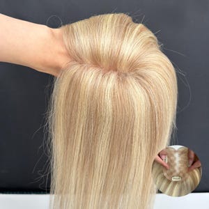 May include: Blonde hair topper with highlights, held up to show the hair's texture and color. The hairpiece appears to be made of real hair, with a natural-looking blend of blonde and lighter streaks. The topper has clips for easy attachment.