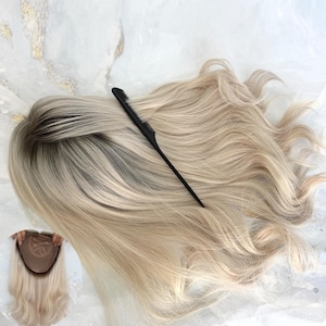 Puede incluir: Una peluca rubia con raíces oscuras y cabello largo y ondulado. Un peine negro descansa sobre la peluca. La peluca se muestra con un primer plano de la gorra y el cabello.