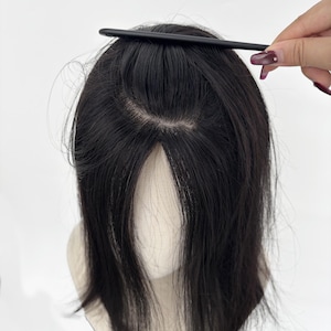 Puede incluir: Una peluca negra con cabello oscuro, peinada con un peine. La peluca se coloca sobre una cabeza de maniquí. El cabello está partido por la mitad, con una línea de cabello de aspecto natural.