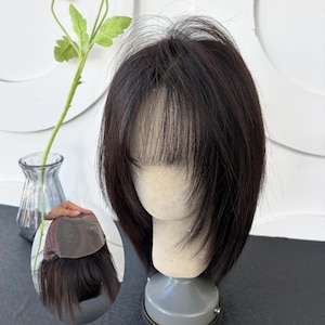 May include: A dark brown hair topper with wispy bangs and layered ends, styled on a mannequin head. An inset image shows the underside of the hair topper, revealing a brown mesh base with clips for attachment to natural hair.