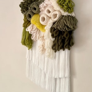 May include: A textured wall hanging featuring a variety of yarn colors and textures. The piece includes shades of green, yellow, white, and beige, with a fringe at the bottom. The wall hanging is mounted on a wooden dowel.