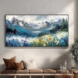 Könnte beinhalten: Ein großes Landschaftsgemälde zeigt eine Bergkette mit schneebedeckten Gipfeln unter einem bewölkten Himmel. Der Vordergrund zeigt ein Feld bunter Wildblumen in Blau-, Gelb- und Weißtönen. Das Kunstwerk ist gerahmt und hängt an einer strukturierten weißen Wand.
