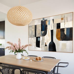 May include: A dining room scene featuring a light wood table with black chairs. A large abstract painting with black, white, and tan shapes hangs on the wall. A woven sphere pendant light hangs above the table, and a vase of pink flowers sits on the table.
