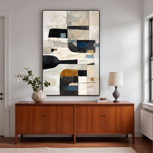 May include: Abstract art piece in a black frame. The artwork features a geometric design with shapes in black, white, beige, and touches of blue and brown. The painting hangs above a wooden cabinet with a lamp and vase.