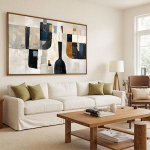 May include: An abstract painting in a wooden frame hangs above a white sofa with green pillows. The artwork features a mix of shapes and colors, including black, white, and brown. A wooden coffee table and a floor lamp complete the living room setting.