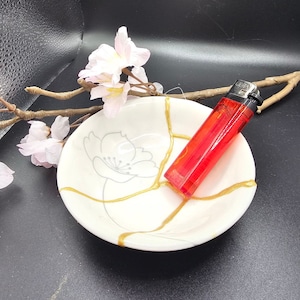 Puede incluir: Un plato de cerámica blanca con esmalte craquelado dorado y un diseño floral. Un encendedor rojo está sentado en el plato.