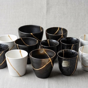 Może przedstawiać: Kolekcja czarnych i białych ceramicznych kubków i misek, z których każdy ma złote linie naprawy kintsugi. Naczynia różnią się rozmiarem i kształtem, niektóre mają teksturowane powierzchnie. Układ znajduje się na neutralnym tle.