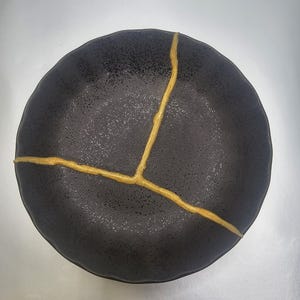 Könnte beinhalten: Ein runder, schwarzer Keramikteller mit goldfarbenen Kintsugi-Reparaturlinien. Der Teller hat eine strukturierte Oberfläche und eine leicht unregelmäßige Form. Die goldenen Linien heben die Risse hervor und schaffen ein einzigartiges Design. Ideal zum Servieren von Speisen oder als Ausstellungsstück.