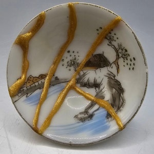 May include: A white porcelain saucer with a blue and black landscape scene painted on it. The saucer is cracked and the cracks are filled with gold paint.