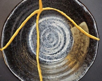 May include: Black ceramic bowl with a gold crackle glaze. The bowl is circular in shape and has a slightly raised rim.