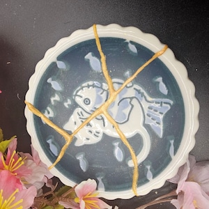 May include: A small, round, blue and white ceramic dish with a gold crackle design. The dish features a stylized fish design and is perfect for holding small items.