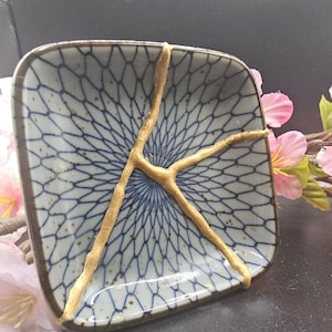 Puede incluir: Un pequeño plato cuadrado de cerámica con un patrón geométrico azul y blanco. El plato tiene un diseño craquelado de esmalte dorado.