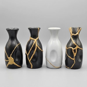 May include: Four ceramic vases displayed in a row. Two are black with gold-coloured crack patterns, one is white with a gold crack, and the last is black with gold accents. The vases have a narrow neck and a rounded body.