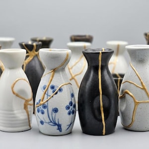 May include: A selection of ceramic sake bottles, demonstrating the Japanese art of Kintsugi. The bottles are in various colours, including black, white, and speckled grey, with gold-coloured repair lines. Some bottles feature blue floral designs.