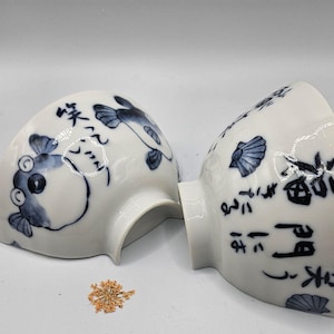May include: Two white ceramic bowls with blue and white designs. One bowl features a pufferfish and the other features a seashell and Japanese characters.
