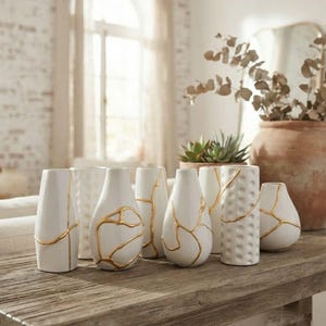 May include: A collection of eight white ceramic vases arranged on a wooden surface. Each vase features unique gold-colored lines resembling kintsugi repair, with some vases displaying smooth surfaces and others showing a textured, dimpled pattern.