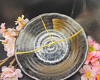 May include: A black and blue ceramic bowl with gold detailing, demonstrating the Japanese art of Kintsugi. The bowl is circular with a textured surface and gold lines. Pink flowers are arranged around the bowl.