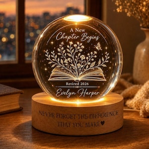 May include: A spherical glass night light on a round wooden base. The glass features an etched open book with floral sprigs, a butterfly, and the text A New Chapter Begins, Retired 2026, and Evelyn Harper. The base reads NEVER FORGET THE DIFFERENCE THAT YOU MAKE.