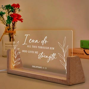 May include: A rectangular LED light with a wooden base displays the text "I can do all this through him who gives me strength" in white script. The light has a warm glow and is decorated with floral accents.