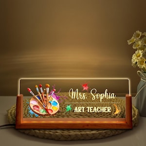 May include: A personalized art teacher desk lamp with a wooden base. The lamp features a clear acrylic panel with the name "Mrs. Sophia" and the words "ART TEACHER" illuminated in a warm glow. A palette with paintbrushes and colorful paint splatters are also displayed.