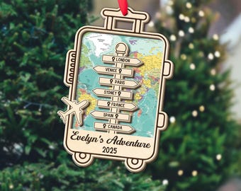 May include: Wooden suitcase-shaped Christmas ornament with a world map background. Features directional signs pointing to London, Venice, Paris, Sydney, France, Spain, and Canada. Includes a small airplane cutout and the text "Evelyn's Adventure 2025".