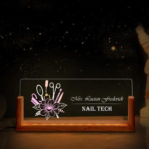 May include: A lighted acrylic sign with a wooden base, featuring a floral design and nail tools. The sign displays the name "Mrs. Lucian Frederick" and the title "NAIL TECH" in elegant script. The background is a dark, starry night.