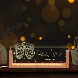 May include: A lighted desk sign with a wooden base and a clear acrylic panel. The panel features an optometrist's instrument design and the name "Hailey Smith, Optometrist". The sign is illuminated with warm white lights.