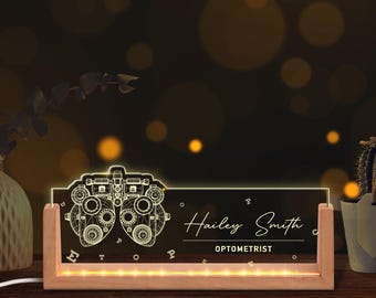May include: A lighted desk sign with a wooden base and a clear acrylic panel. The panel features an optometrist's instrument design and the name "Hailey Smith, Optometrist". The sign is illuminated with warm white lights.