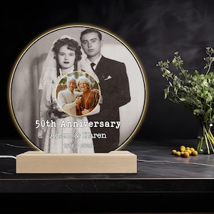 May include: A round, illuminated photo frame with a black and white image of a couple, and a smaller color photo of the same couple. The text "50th Anniversary" and names are displayed. The frame sits on a wooden base.