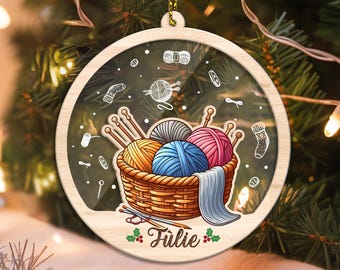 May include: Round wooden Christmas ornament with a clear center, featuring a basket of yarn balls in orange, blue, and pink. Knitting needles and a blue scarf are also in the basket. The name "Julie" is written below the basket.