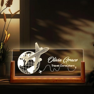May include: A lighted desk sign with a wooden base. The clear acrylic panel features a globe and airplane design, with the name "Olivia Grace" and the title "Travel Consultant" in white script. The sign is illuminated with a warm glow.