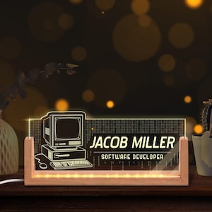 May include: A personalized desk nameplate with a retro computer graphic. The clear acrylic panel displays the name "JACOB MILLER" and the title "SOFTWARE DEVELOPER" in white. The nameplate is set in a wooden base with embedded lights.