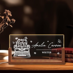 May include: An illuminated desk sign with a wooden base. The black acrylic panel features a white illustration of a typewriter with flowers, butterflies, and the name "Amelia Carson, Writer."