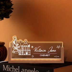 May include: A lighted desk nameplate with a wooden base. The clear acrylic panel features a microscope, test tubes, and molecular structures. The name "Victoria Jone" and the title "CHEMIST" are displayed in white script.