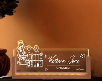 May include: A lighted desk nameplate with a wooden base. The clear acrylic panel features a microscope, test tubes, and molecular structures. The name "Victoria Jone" and the title "CHEMIST" are displayed in white script.