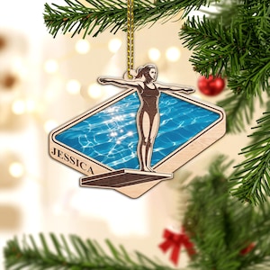 May include: Wooden Christmas ornament featuring a diver in a swimsuit, arms outstretched, standing on a diving board. The background shows a pool of water. The name "JESSICA" is printed on the ornament. The ornament is hanging from a gold string.