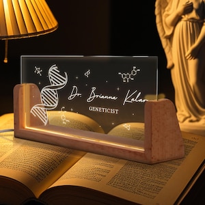 May include: A desk lamp featuring a wooden base and a clear acrylic panel. The panel displays a DNA strand, molecular structures, and the name "Dr. Brianna Kalar, Geneticist." The lamp emits a warm, soft light, creating a gentle glow.