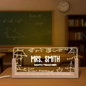 May include: A personalized desk nameplate for a math teacher. The illuminated acrylic sign, set in a wooden base, displays "MRS. SMITH" and "MATH TEACHER" in white, along with mathematical formulas and geometric shapes. The sign is lit with a warm glow.