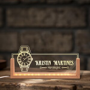 May include: A personalized desk nameplate with a watch design and the name "KRISTIN MARTINES, Horologist" in elegant font. The clear acrylic panel is illuminated by warm white LED lights, set in a wooden base.