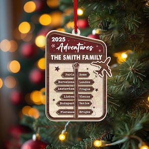 May include: Wooden Christmas ornament with a travel theme. The ornament is rectangular with a light brown background and a dark brown border. It features the text "2025 Adventures The Smith Family" and a list of cities. A small airplane cutout is attached.