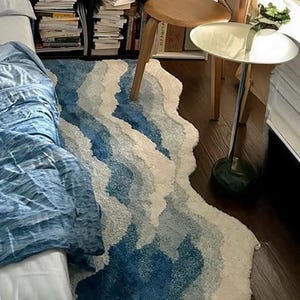 May include: A bedroom with a blue bedspread and a blue and white rug. The wall is adorned with framed pictures and plants. A wooden chair and small table are positioned near a window, creating a tranquil space.