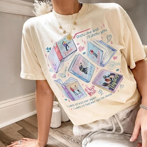 Könnte beinhalten: Ein cremefarbenes Kurzarm-T-Shirt mit einem Grafikdruck von vier offenen Büchern mit den Titeln The Deal, The Mistake, The Score und The Legacy von Elle Kennedy. Das Design enthält kleine Herzen, Sterne und den Text Show me and I'll show you.