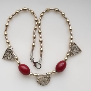 May include: Silver-toned necklace with a mix of small, elongated beads and larger, oval red beads. The necklace features triangular pendants with intricate designs and a decorative clasp.