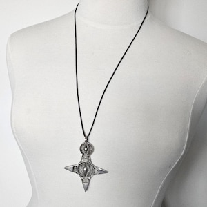 May include: A silver-colored pendant necklace with a starburst design. The pendant is suspended from a black cord. The necklace is displayed on a white mannequin.