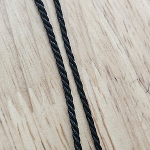May include: Close-up of two strands of black twisted cord, likely for jewelry or crafts. The cord has a textured, rope-like appearance. The background is a light-colored wooden surface.