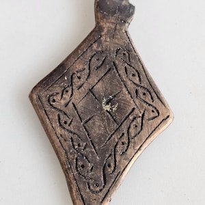 May include: A bronze-coloured pendant with a diamond shape and a loop at the top. The pendant features an engraved design with a central square and decorative borders. The metal has a slightly aged appearance.