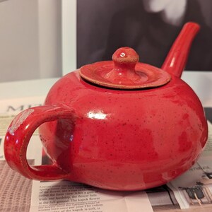 May include: A red ceramic teapot with a speckled glaze. The teapot has a rounded body and a short spout. The lid is also red and has a small knob on top.