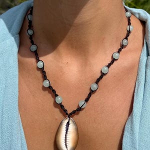 May include: A handmade necklace featuring a large, tan and white seashell pendant. The necklace has a black cord with light blue beads spaced along the length. The necklace is a bohemian style.
