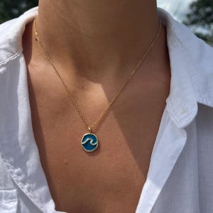 May include: Gold-toned necklace with a circular pendant featuring a blue wave design. The pendant is outlined in gold and hangs from a delicate chain. The necklace is worn against a white shirt.